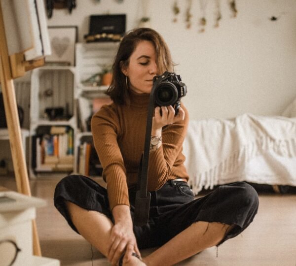 woman sitting on brown wooden floor while holding black DSLR camera in room